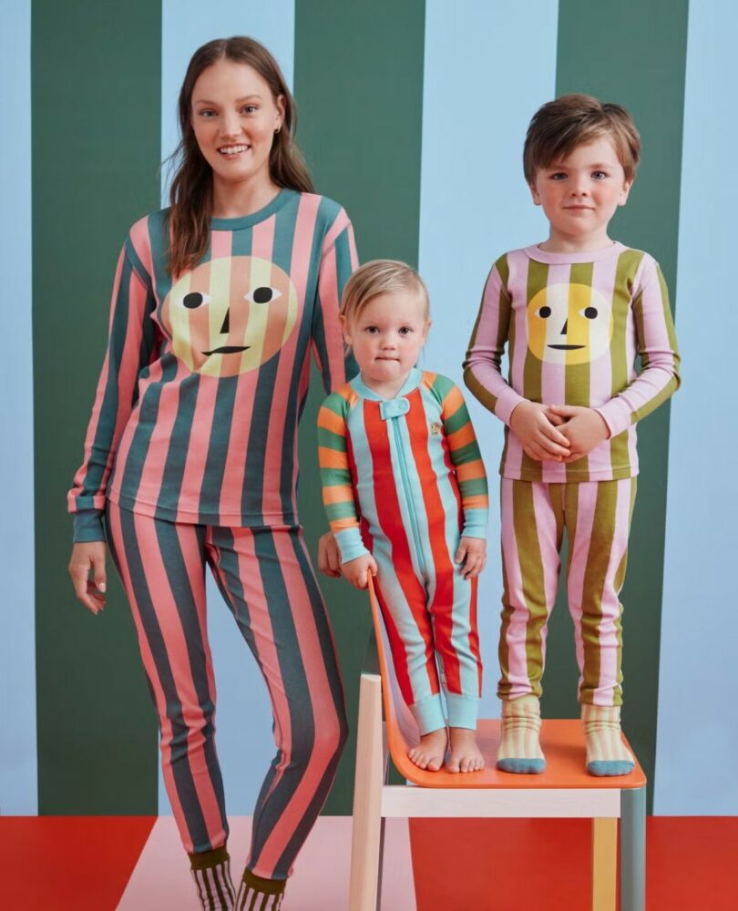 A woman and two children pose in colorful, striped pajamas with face designs, standing and sitting in front of a striped background