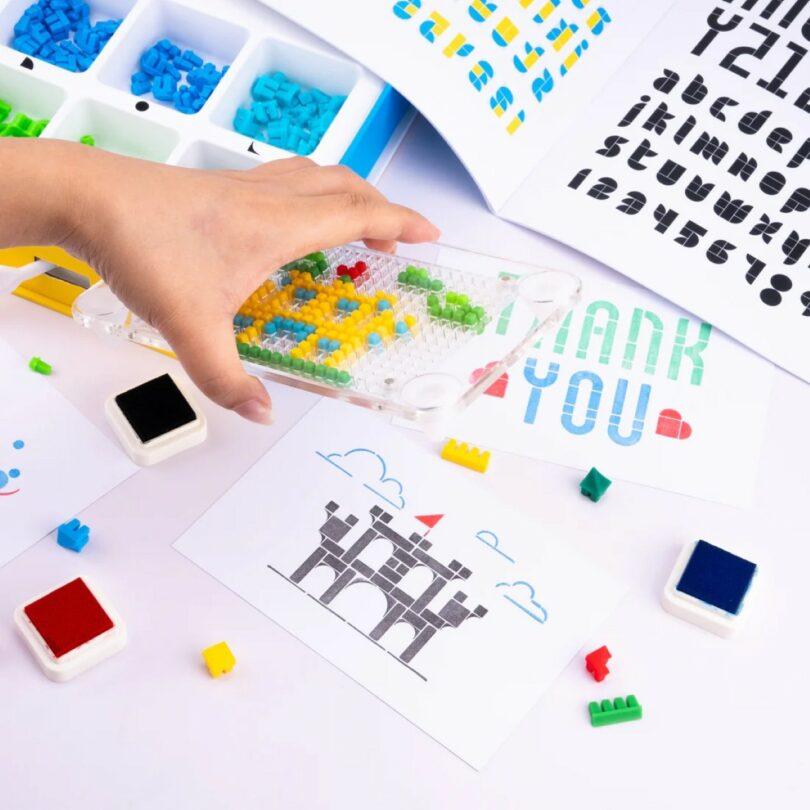 A hand arranges small colored pegs on a clear pegboard, surrounded by ink stamps, peg pieces, and printed cards with letters and a castle illustration