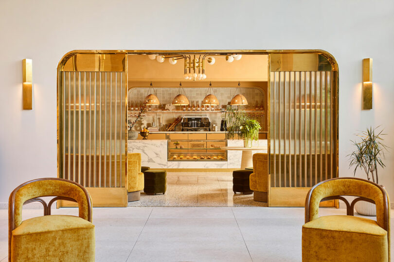 A modern cafe interior with gold accents, plush yellow chairs, marble countertops, and warm lighting, framed by fluted glass sliding doors.