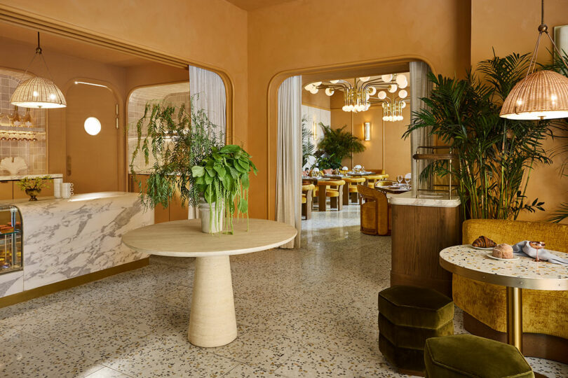 Modern cafe interior with warm tan walls, terrazzo floors, marble table, greenery, velvet seating, and wicker pendant lights. Dining tables are set in the background.