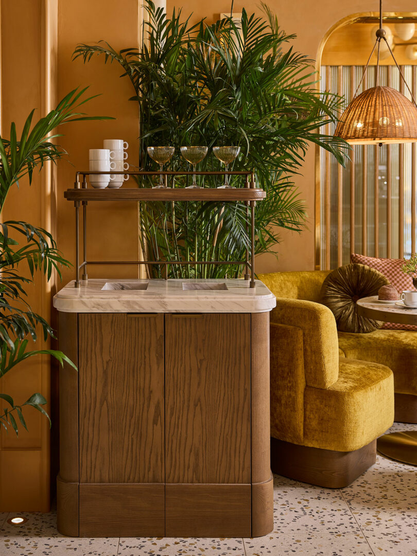 A wooden sideboard with a marble top holds stacked cups and glassware, set against green plants and mustard yellow seating in a warmly lit, stylish interior.