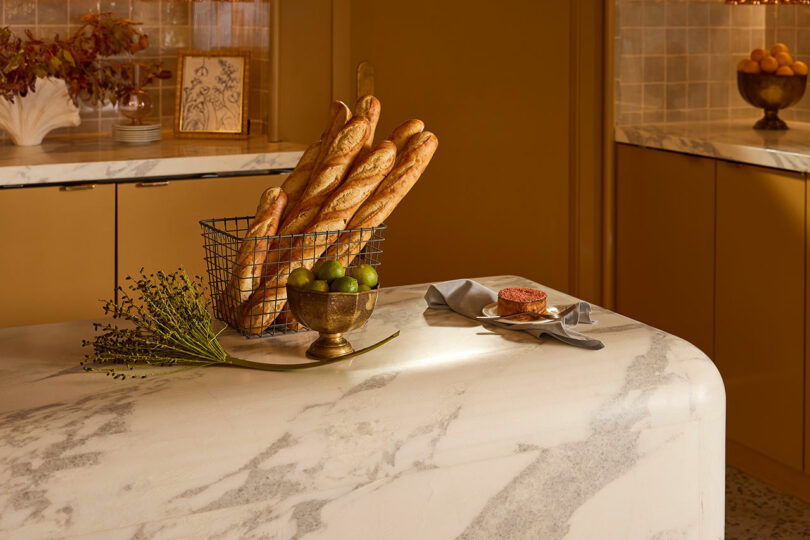 A marble kitchen island with a wire basket of baguettes, a brass bowl of green fruit, a branch of greenery, and a plate with pâté and toast on a napkin.