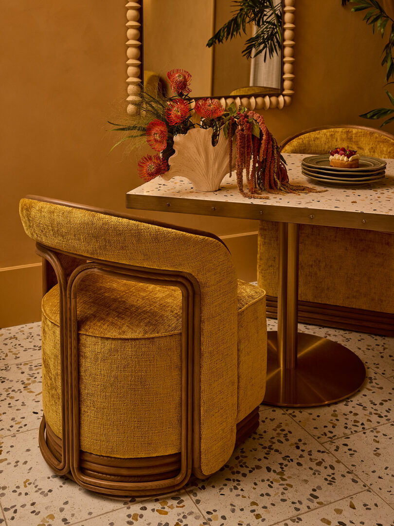 A gold upholstered chair sits at a terrazzo table with gold base, decorated with a sculptural vase of flowers; a mirror and green plant are visible in the background.
