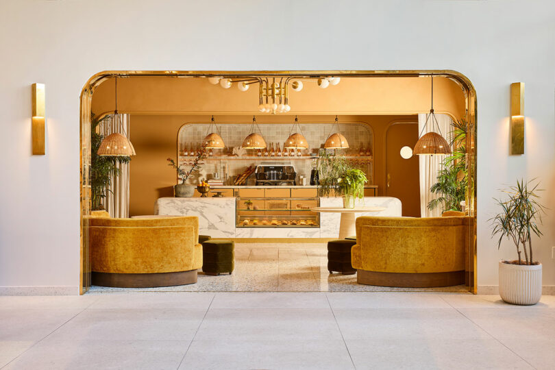 A modern café with gold velvet seating, marble counter, woven pendant lights, and potted plants, featuring warm beige and gold tones.