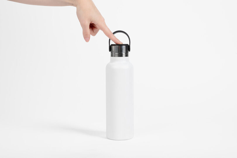 A hand presses the black lid of a plain white reusable water bottle against a white background.