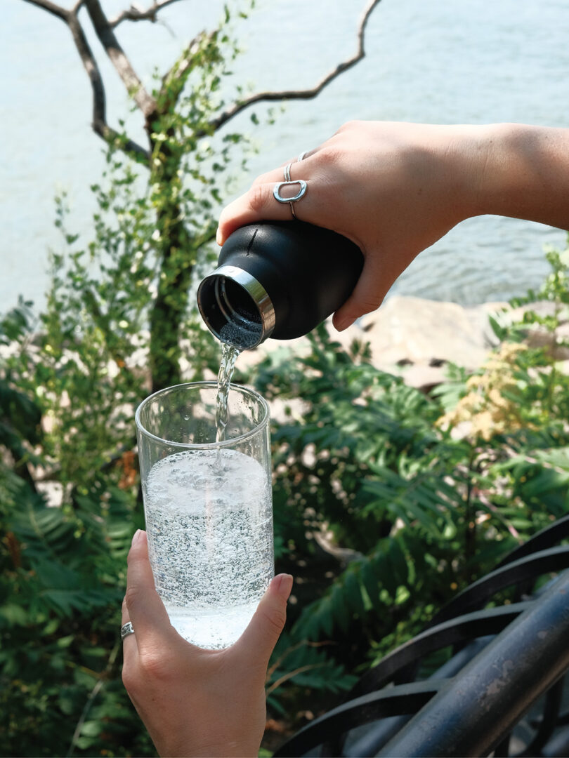 A person pours water from a black bottle into a glass outdoors, with greenery and a body of water in the background.