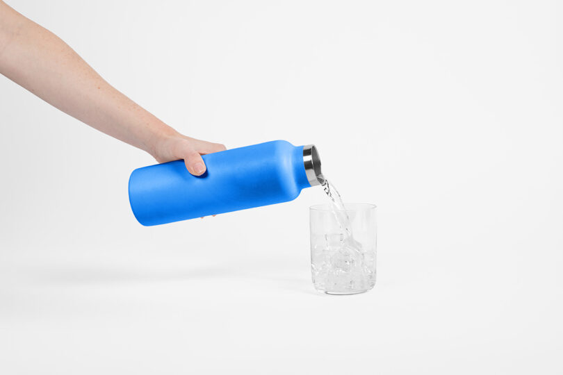 A hand pours water from a blue bottle into a clear glass filled with ice, against a plain white background.