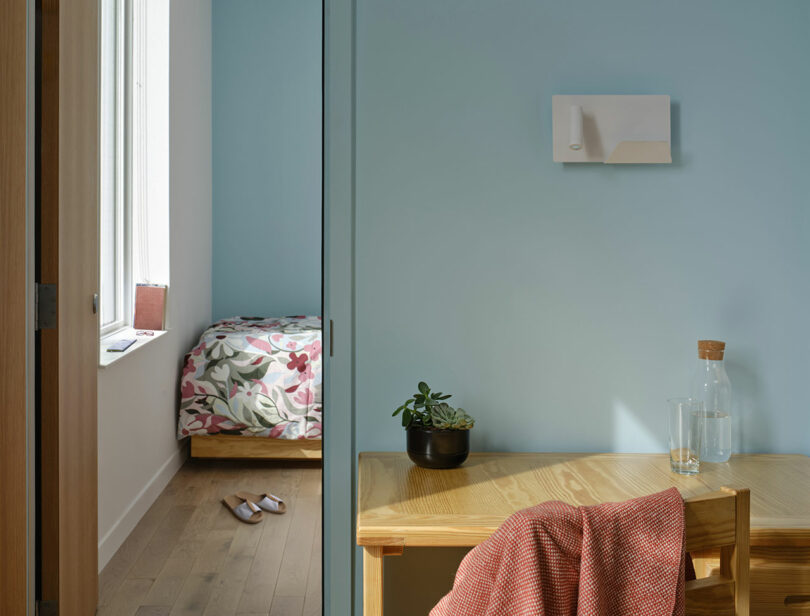 A tidy bedroom with a floral bedspread and slippers on a wooden floor, adjacent to a workspace with a wooden desk, a plant, glass water bottle, and a coral-patterned cloth on a chair.
