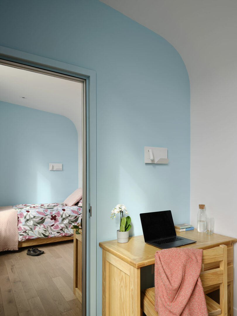 A small wooden desk with a laptop, glass bottle, and an orchid stands near an open door leading to a bedroom with a floral bedspread and blue walls.