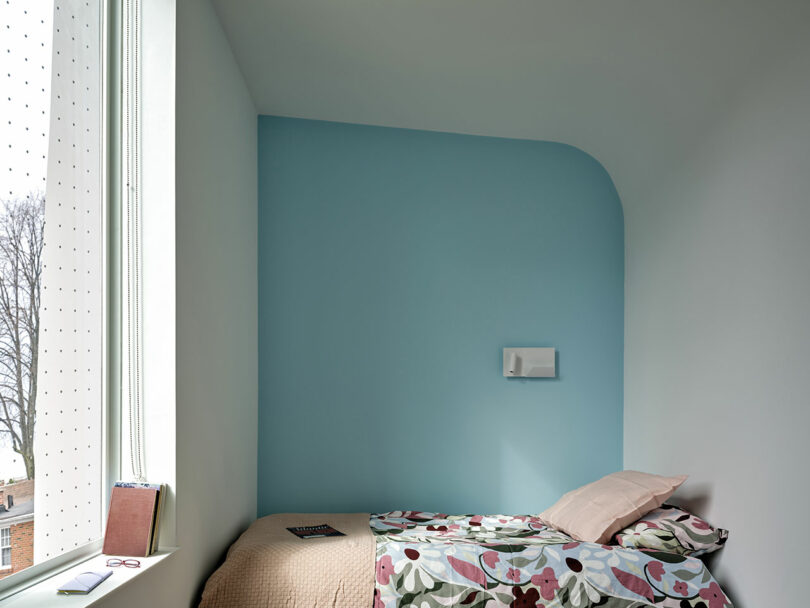 A minimalist bedroom with a single bed, floral bedding, blue accent wall, and a large window with a book and notebook on the windowsill.