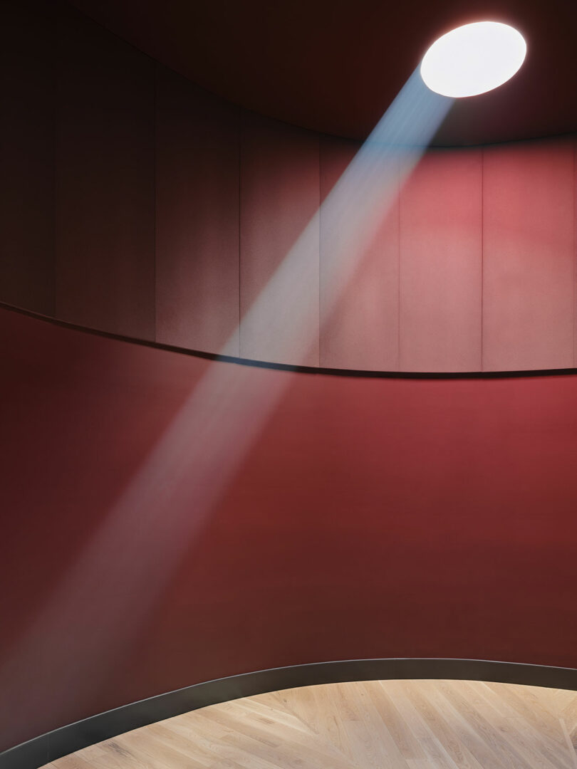 A circular skylight casts a beam of light onto the wooden floor in a room with curved, matte red walls.