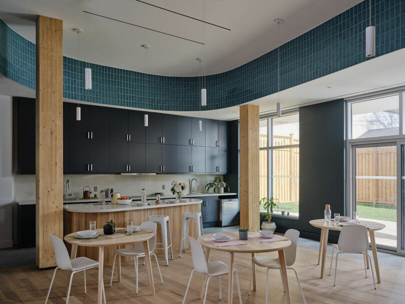 Modern kitchen and dining area with light wood flooring, round tables set for meals, dark cabinets, and a curved blue tile feature above the workspace. Large windows let in natural light.