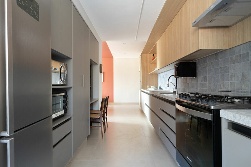Modern galley kitchen with gray and wood cabinets, stainless steel appliances, a gas stove, light tile floor, and a coral accent wall at the far end.