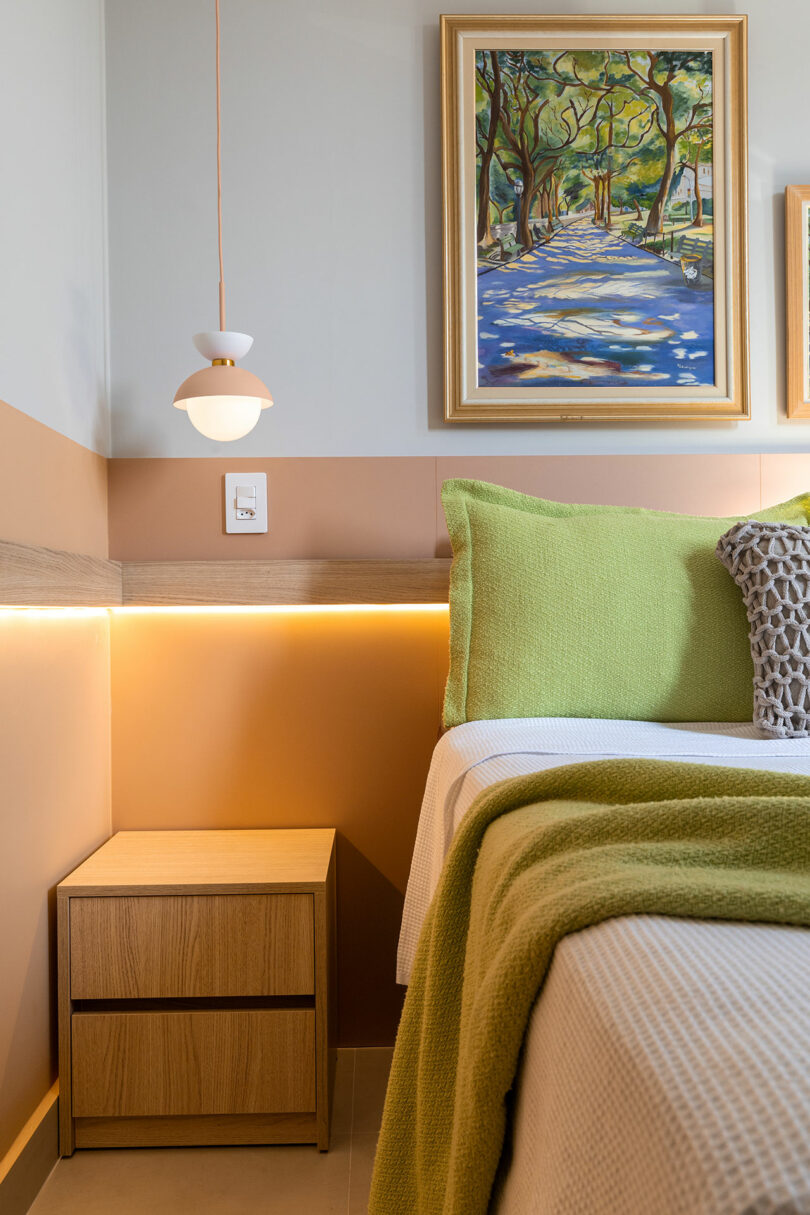 A modern bedroom corner with a wooden nightstand, pendant light, framed painting, and a bed with green and beige bedding.