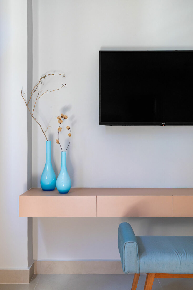 A wall-mounted TV above a minimalist floating shelf with two blue vases holding dried branches and flowers, next to a light blue upholstered chair.