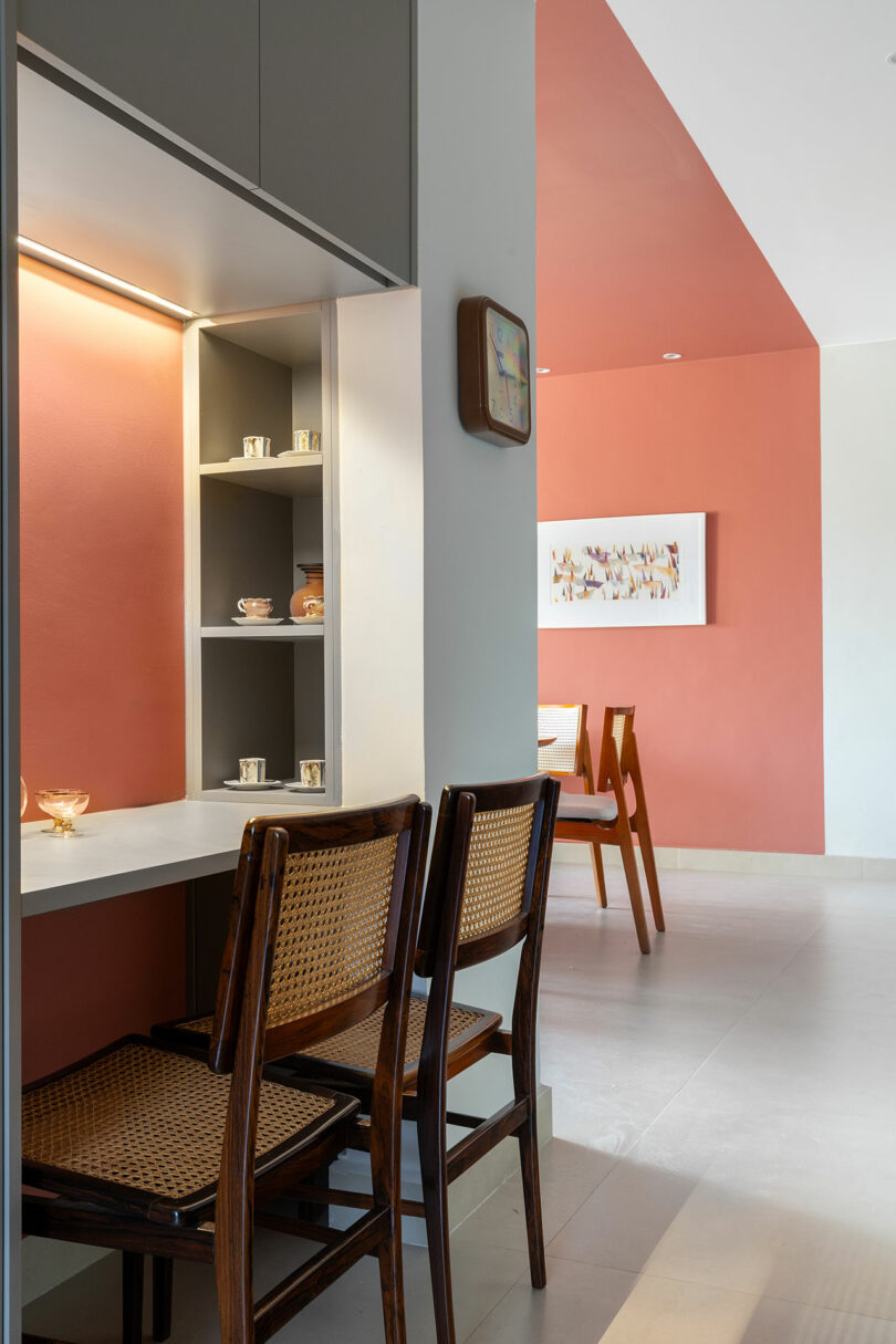Modern dining area with two cane-back chairs at a built-in desk, open shelves with cups, coral-colored accent walls, and a wall clock.