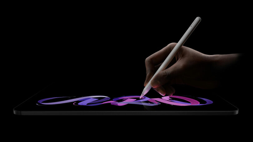 A hand uses a stylus to draw colorful, swirling lines on the screen of an iPad Pro against a black background.