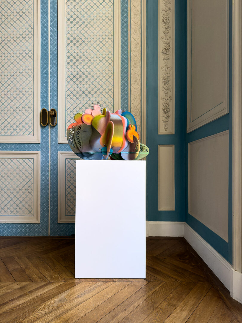 A colorful abstract sculpture sits on a white pedestal beside an iPad Pro in a room with blue and cream patterned walls and a wooden floor.