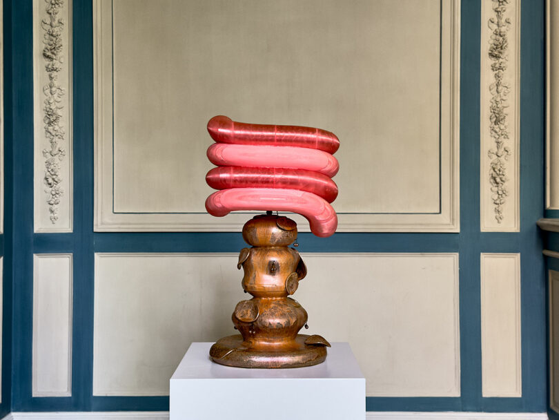 A sculpture featuring a metallic, ornate base topped with four large, pink, tubular forms sits on a white pedestal beside an iPad Pro, all displayed against a decorative wall.