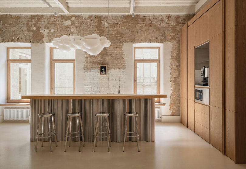 Modern kitchen with a long wooden island, metal bar stools, built-in appliances, exposed brick wall, and large windows. A sculptural white light fixture hangs above the island.