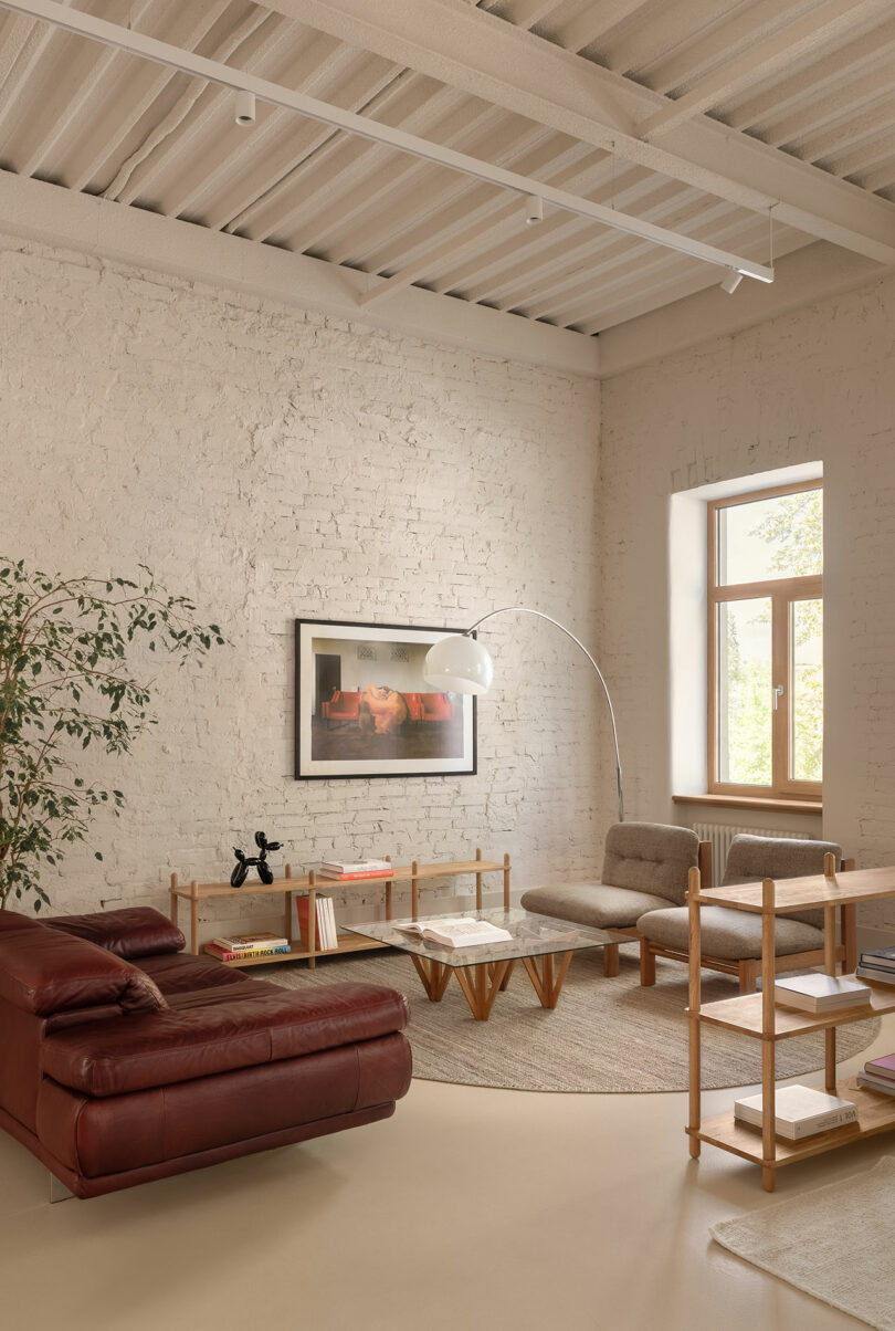 A modern living room with a red leather chaise lounge, beige armchairs, a marble coffee table, shelving unit, potted plant, large window, and framed artwork on a white brick wall.