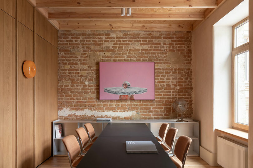 A modern meeting room with a long black table, brown chairs, exposed brick wall, large window, and a pink artwork above a cabinet with a silver globe.