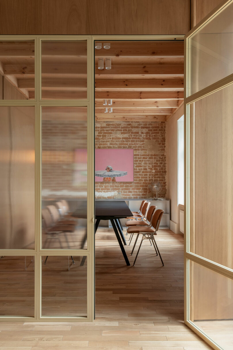 A modern meeting room with a black table, brown chairs, exposed brick wall, wooden ceiling beams, and glass-panel sliding doors.