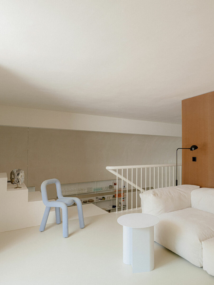Minimalist living space with a white sofa, round white side table, light blue modern chair, beige walls, and a white railing overlooking a lower level with shelves.