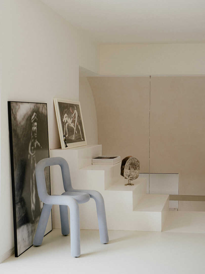 Minimalist interior with a light blue sculptural chair, framed artwork leaning against white steps, and decorative objects on the stairs in a neutral-toned room.