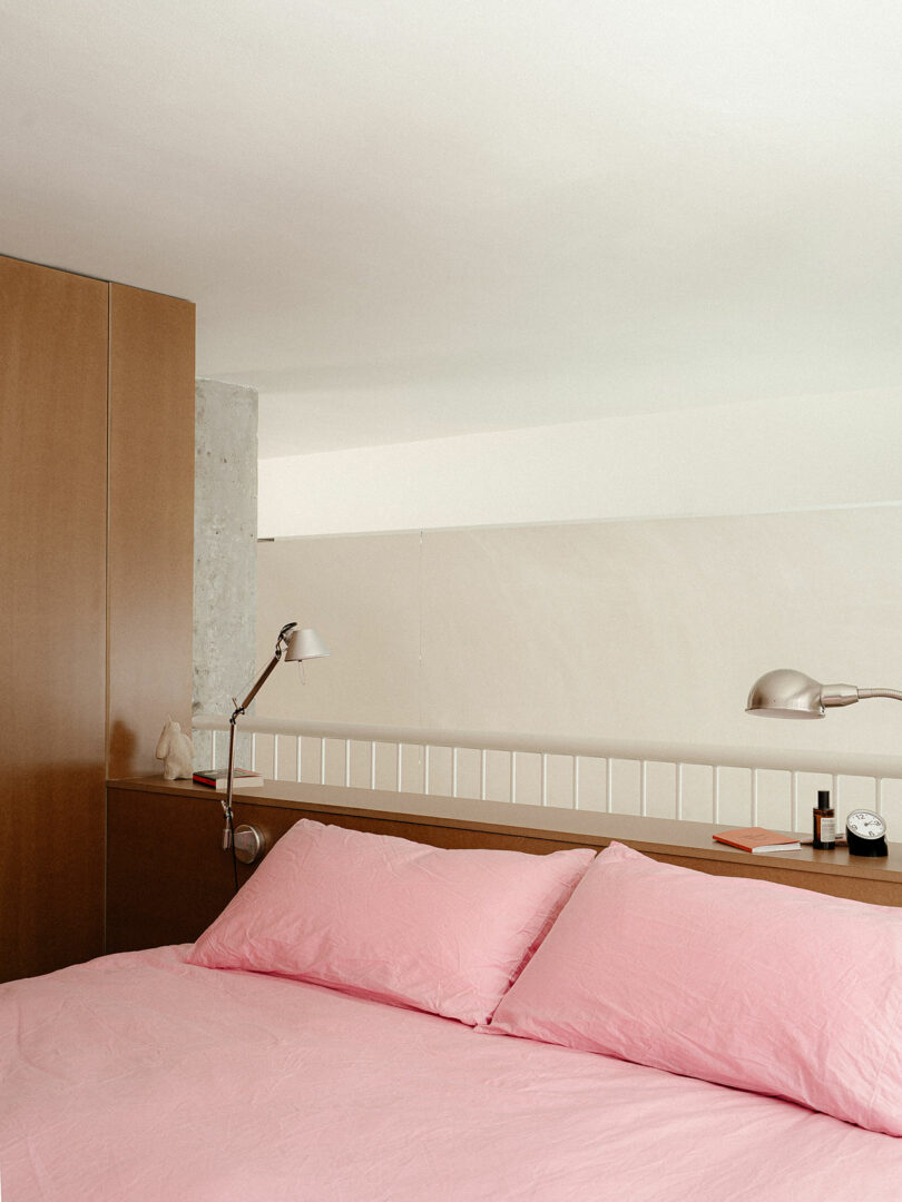 A bed with pink sheets and pillows is placed against a wooden headboard, with two silver lamps, a clock, and small items on the headboard shelf.