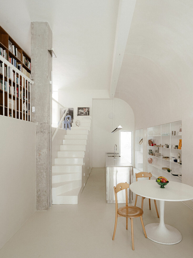 Minimalist kitchen and dining area with white walls, a round table, two wooden chairs, stainless steel kitchen island, open shelving, and a staircase leading to a loft library.