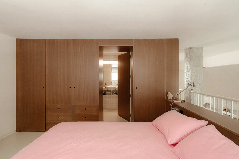 A minimalist bedroom with a pink bedspread, wooden wardrobes, built-in shelving, and an open doorway leading to a bathroom.