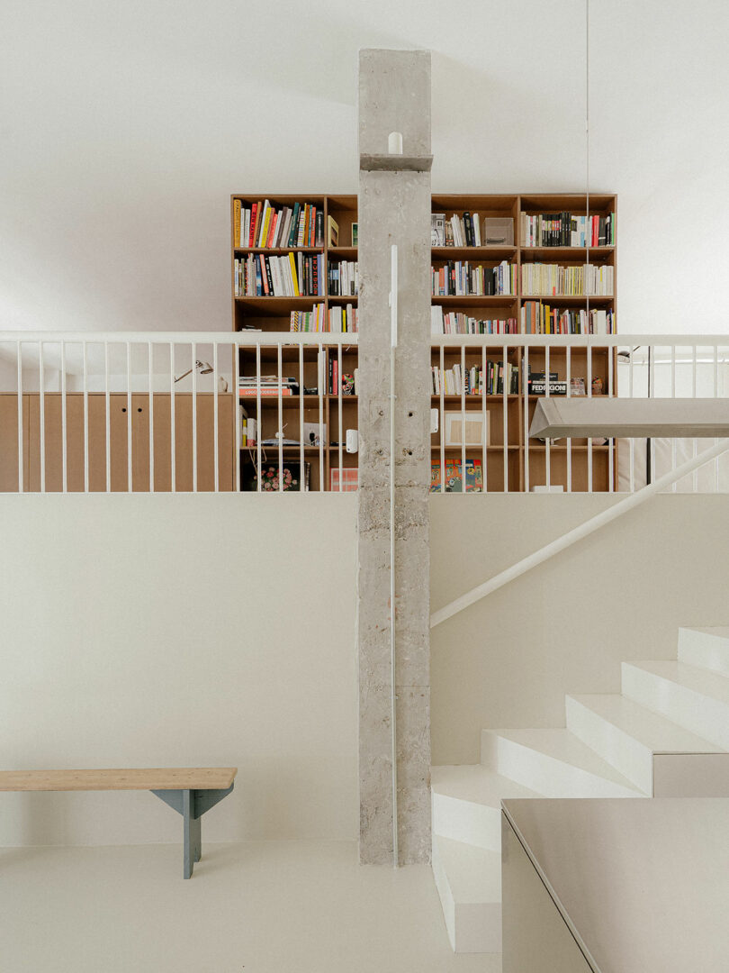 Minimalist interior with white stairs, a concrete pillar, built-in wooden bookshelves filled with books, and a simple wooden bench.