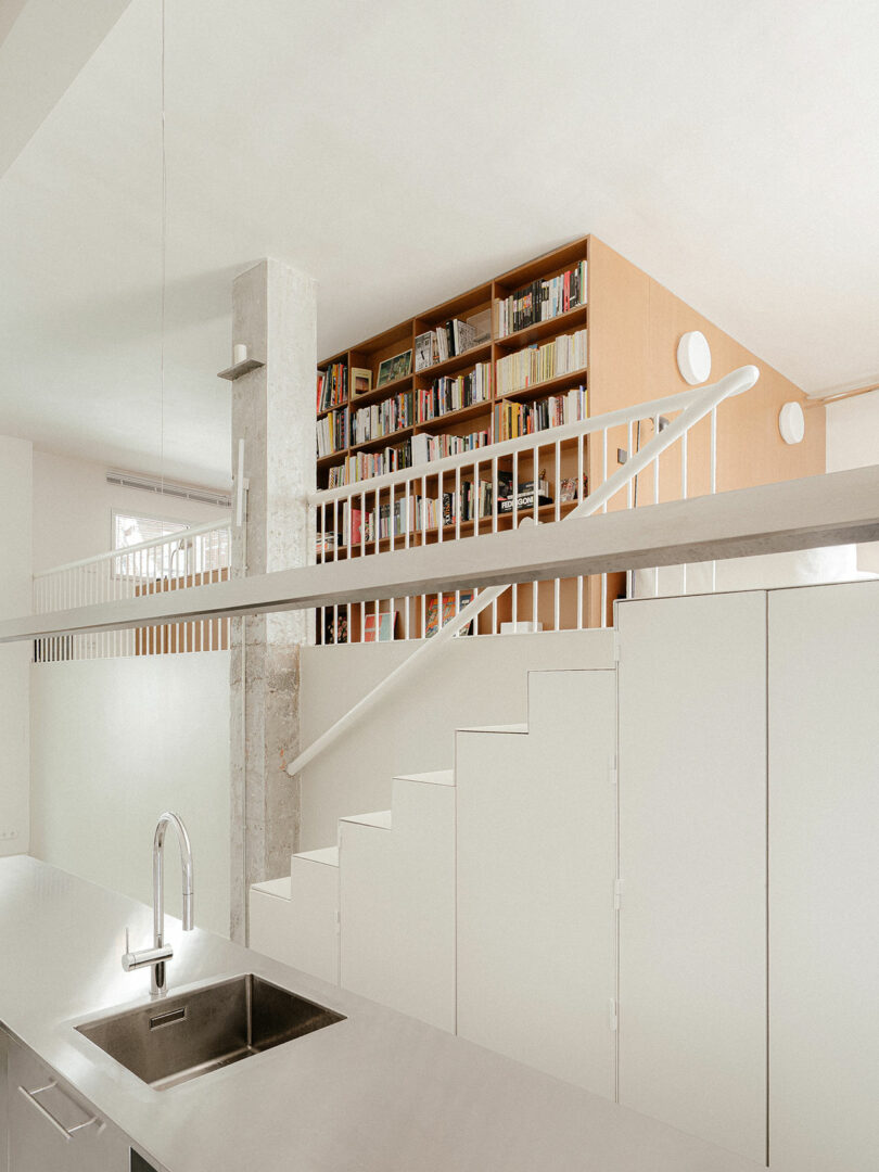 A modern, minimalist interior with white cabinetry, a stainless steel sink, and a staircase leading to a mezzanine with a large bookshelf.