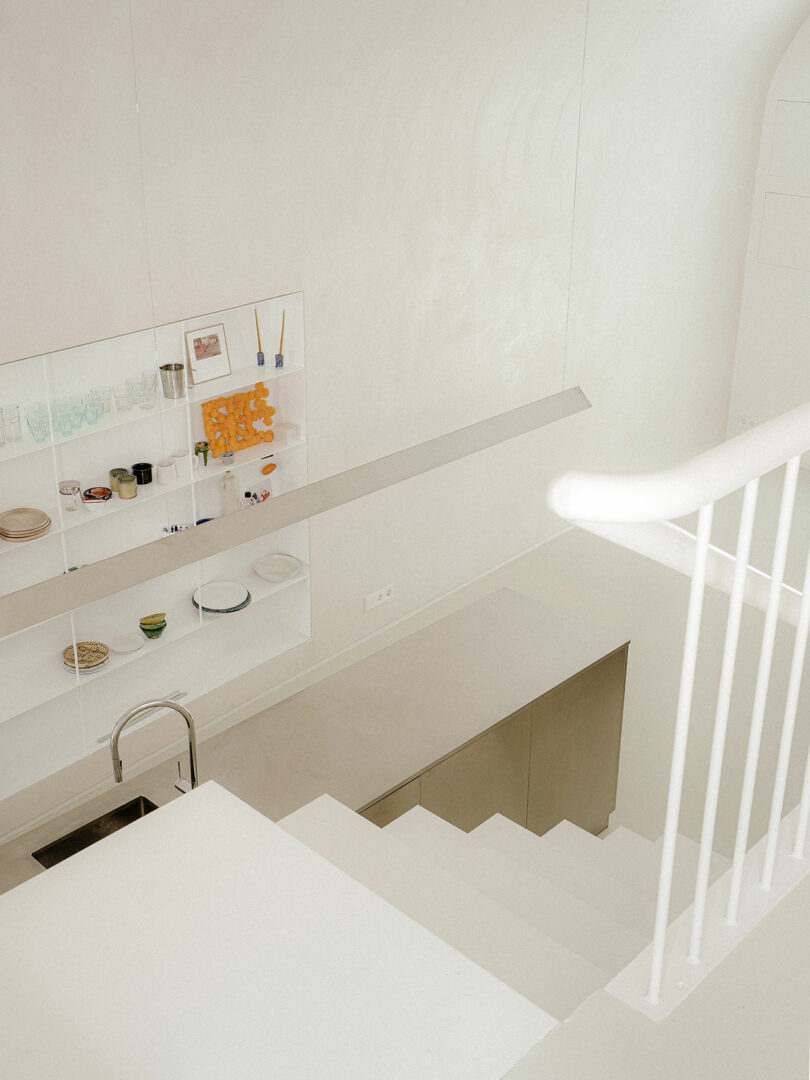 Minimalist kitchen with white walls, open shelving holding dishes and containers, a stainless steel sink, and white staircase railing in the foreground.