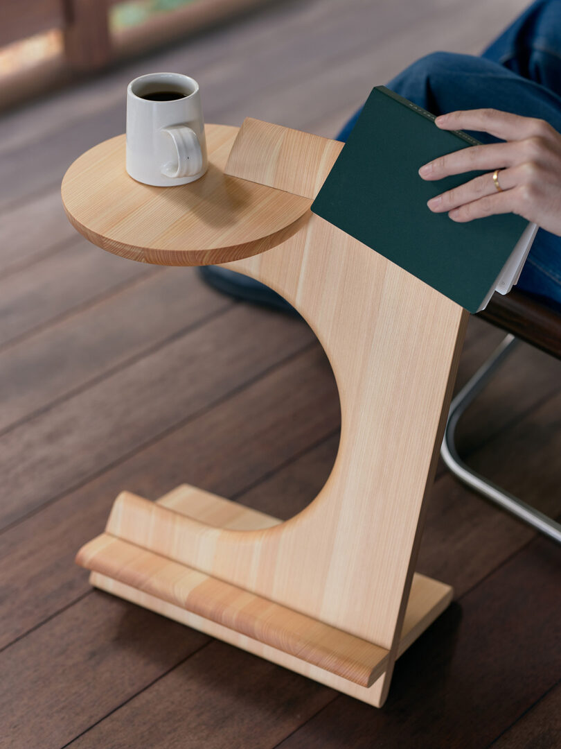 A hand rests a closed green book on a modern wooden side table, which holds a white coffee mug; a person sits nearby on a wooden floor.