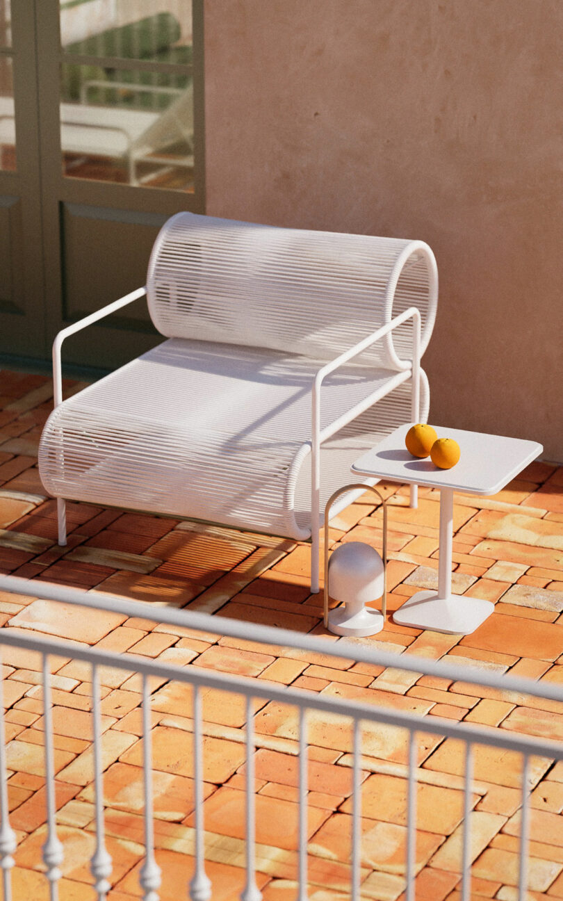 A white modern outdoor chair and side table with two oranges on top sit on a terracotta-tiled patio in bright sunlight.