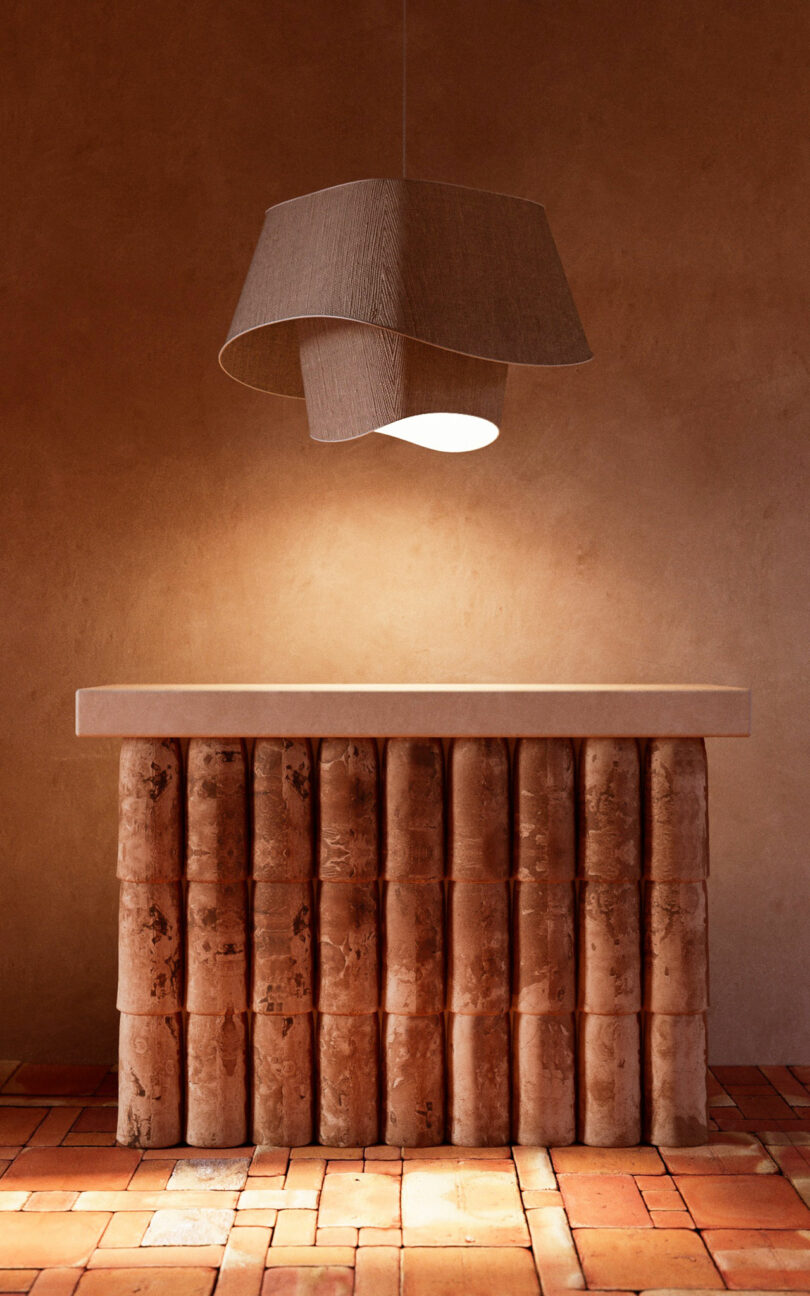 A sculptural wooden table with cylindrical legs stands on terracotta tiles beneath a large, asymmetrical fabric pendant light, set against a warm-toned plaster wall.