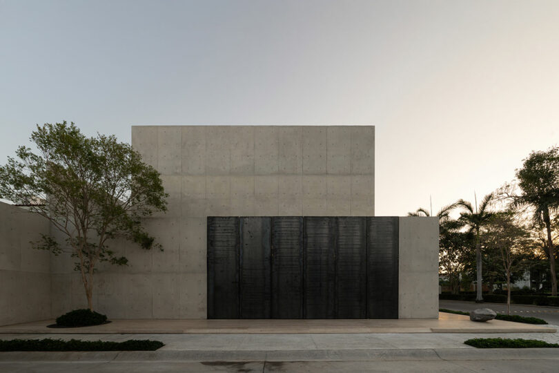 A modern concrete building with a large black metal door, minimal windows, and sparse landscaping with trees and bushes along the sidewalk.
