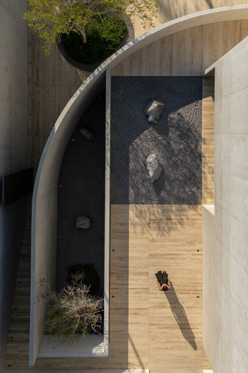 A person in dark clothing stands on a light wood floor in a minimalist courtyard with curved concrete walls, trees, rocks, and gravel patterns, viewed from above.