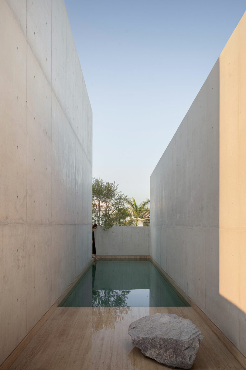 A narrow rectangular pool is set between two tall concrete walls, with a large stone on the tan floor in the foreground and trees visible beyond the pool.