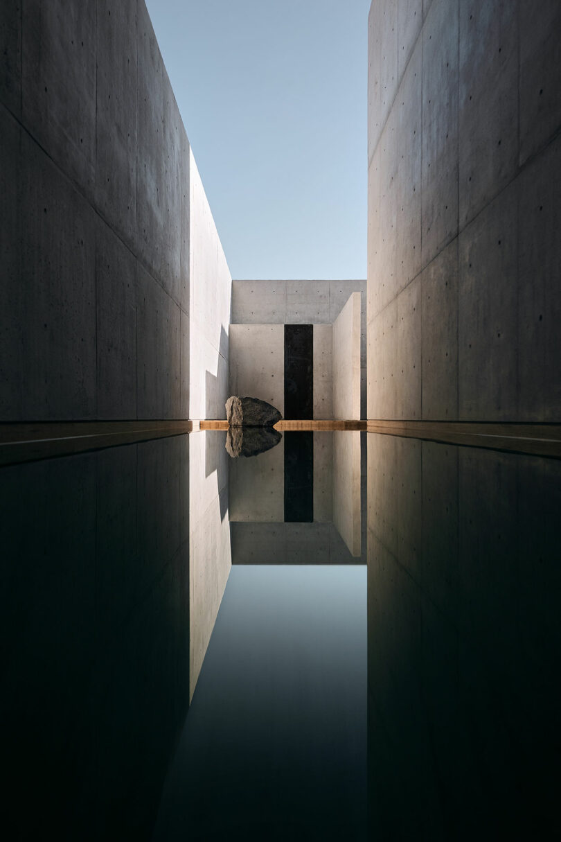 A narrow pool of water reflects minimalist concrete walls and a large rock, creating a symmetrical, serene architectural scene under a clear sky.