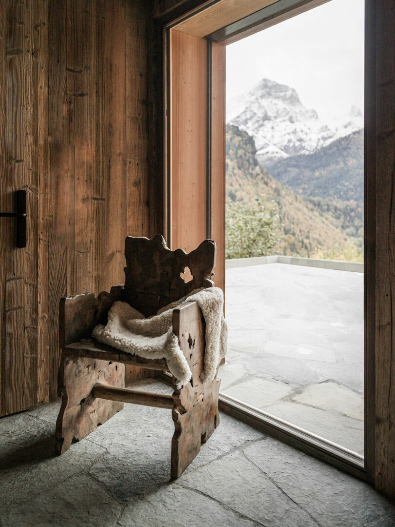 A rustic wooden chair with a sheepskin throw sits by a large window, overlooking a stone patio and snow-capped mountain view.