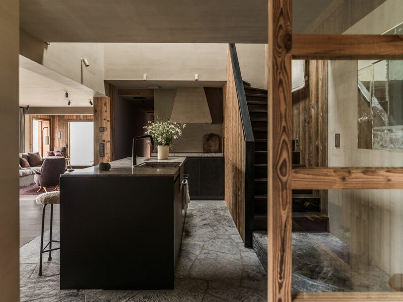 Modern kitchen with a dark island, stone floor, wooden accents, stairs, and a glass partition. A vase with flowers sits on the countertop. Living area visible in the background.