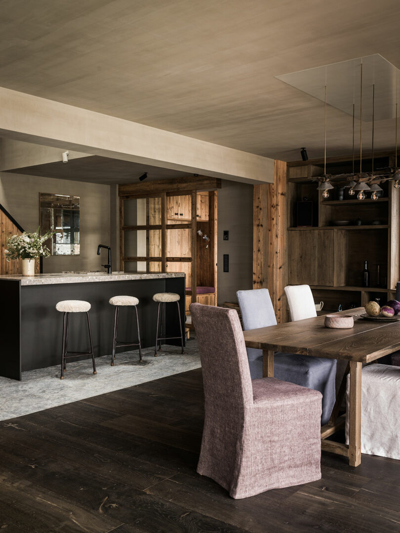 Modern rustic kitchen and dining area with wooden furniture, a black island with three barstools, and a dining table with upholstered chairs on dark wood flooring.