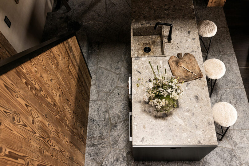 A stone kitchen island with a sink, a black faucet, a rustic wooden cutting board, a vase of flowers, and two round cushioned stools on a stone tile floor.