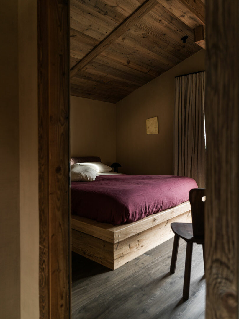 A cozy bedroom with a wooden ceiling and floor, a bed with a burgundy duvet, neutral pillows, a wooden chair, and beige walls with curtains covering a window.