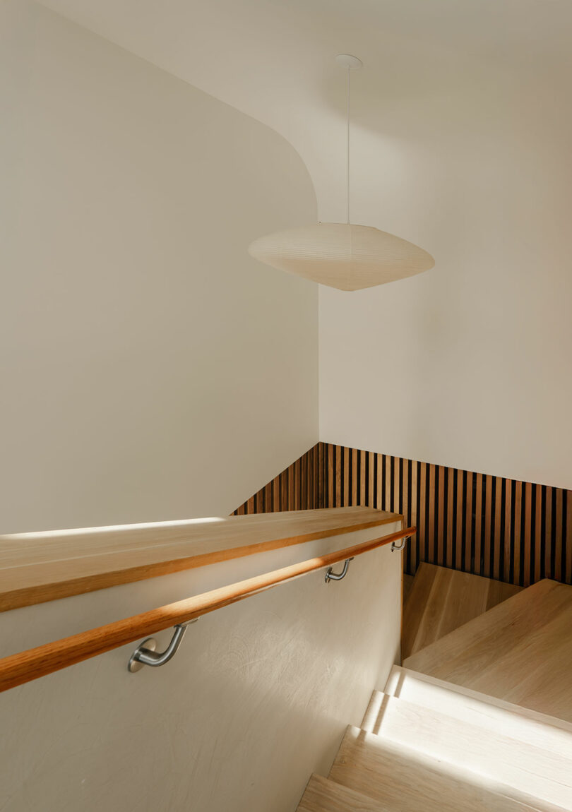 A minimal indoor staircase with light wood steps, a wooden handrail, vertical wood slats on the wall, and a large, round pendant light hanging from the ceiling.