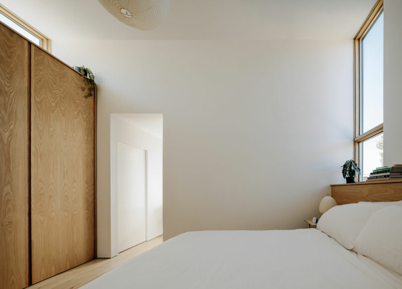Minimalist bedroom with light wood furniture, white bedding, large wardrobe, two windows, and potted plants on the headboard and wardrobe. Natural light fills the space.
