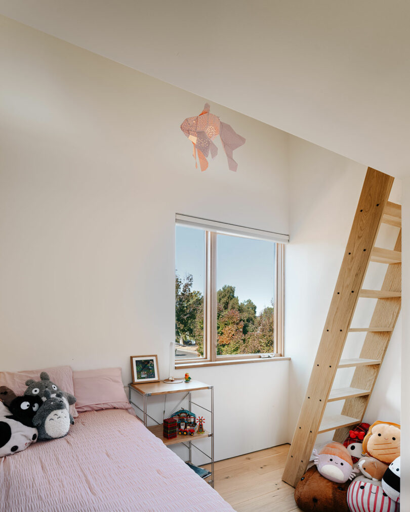 A small bedroom with a loft ladder, a single bed with pink bedding, stuffed toys, a window with a garden view, and a fish-shaped ceiling light.